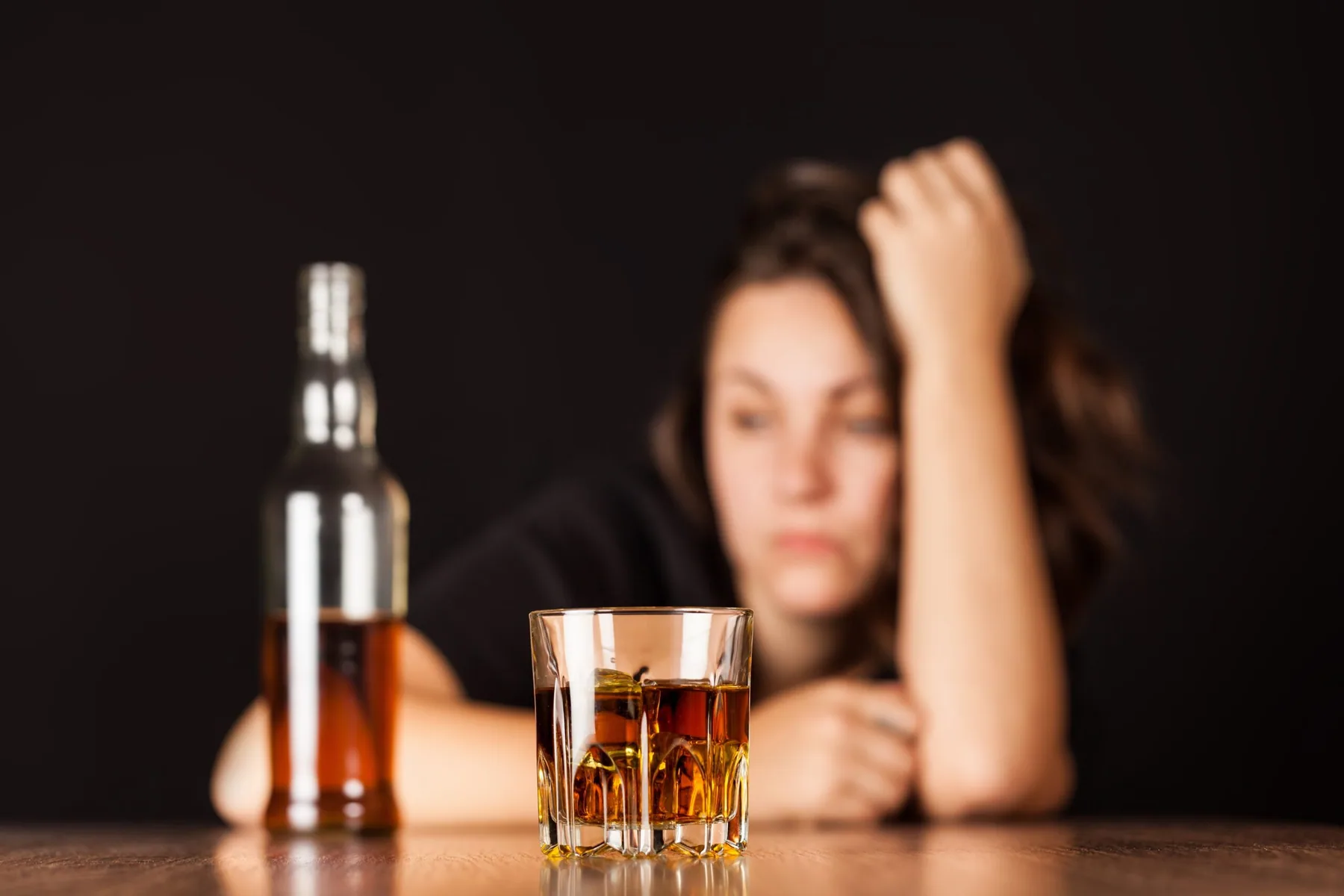 A glass of spirits and a bottle in sharp focus on a wooden table, with a woman resting her head against her arm, out of focus, behind them.