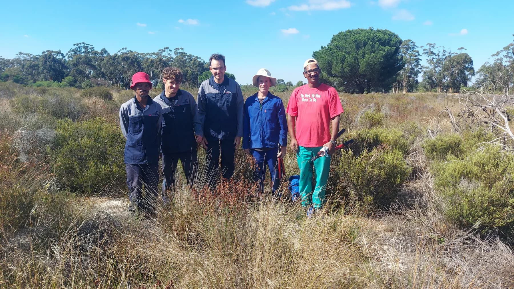 Five members of the Camphill alien clearing team standing together in waist-high fynbos with loppers in hand, cleared veld stretching behind them.