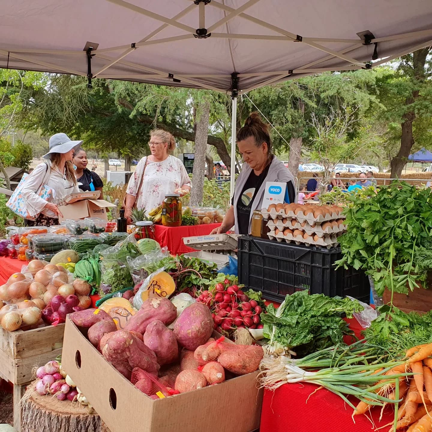 Crates of onions, potatoes, radishes and leafy greens on a produce stall under a white gazebo, with stallholders serving customers behind it