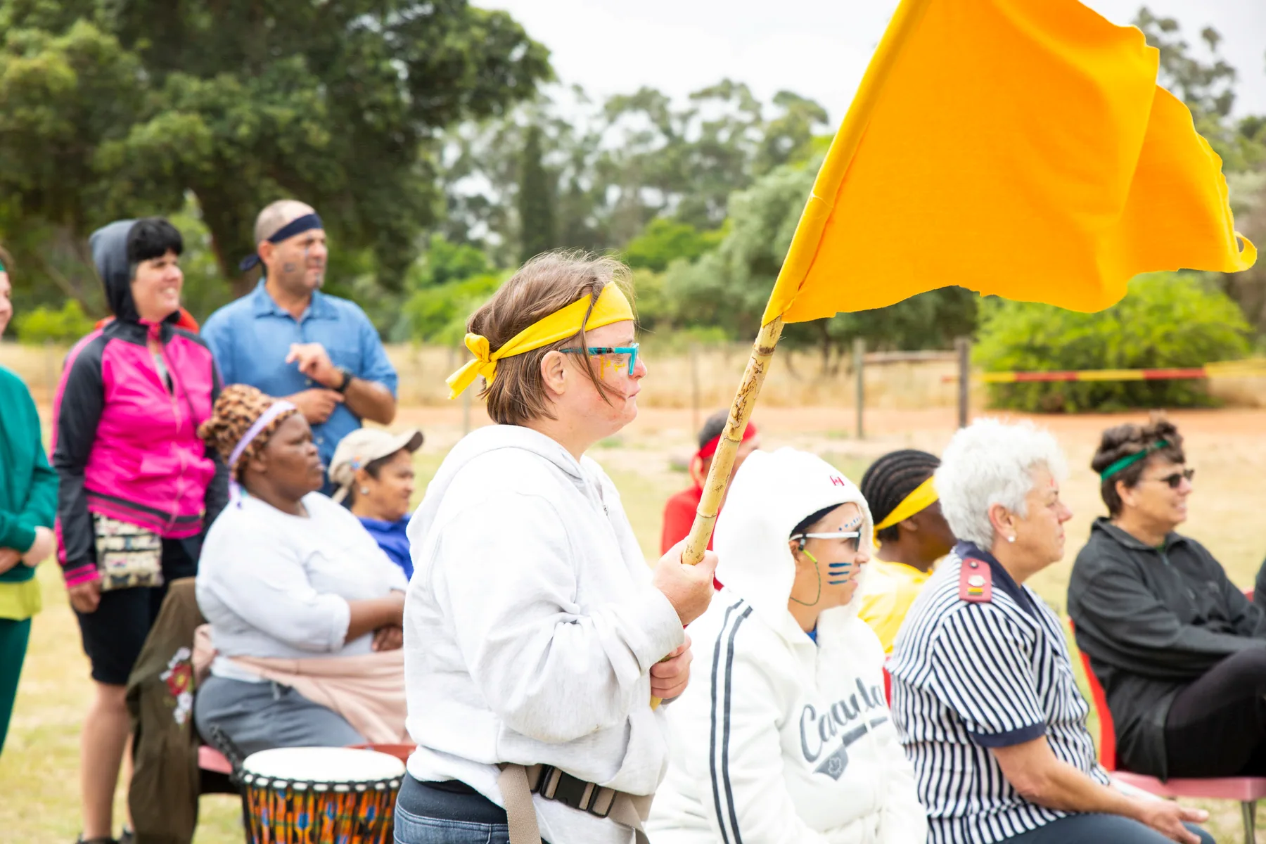 Residents and staff gathered on the village field for a community sports day, one resident carrying a yellow team flag while others sit behind a painted djembe drum.