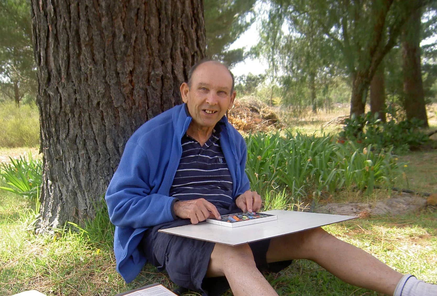 A resident in a blue fleece sitting against a big old tree in the village garden, a board and a tray of colours resting on his knees.