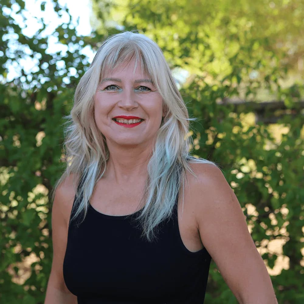 Janine Strumpher, the fundraiser at Camphill Village West Coast, smiling outdoors in front of a leafy green hedge.