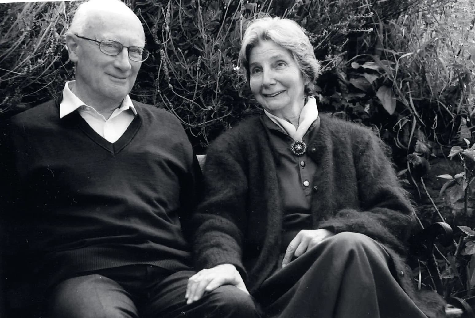 Archive black and white photograph of an older couple sitting together against a bank of foliage, both smiling towards the camera.