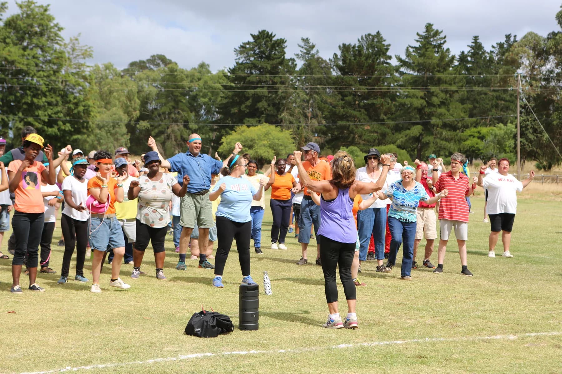Residents, co-workers and staff of Camphill Village warming up together in a wide group on the grass sports field, arms raised mid stretch.