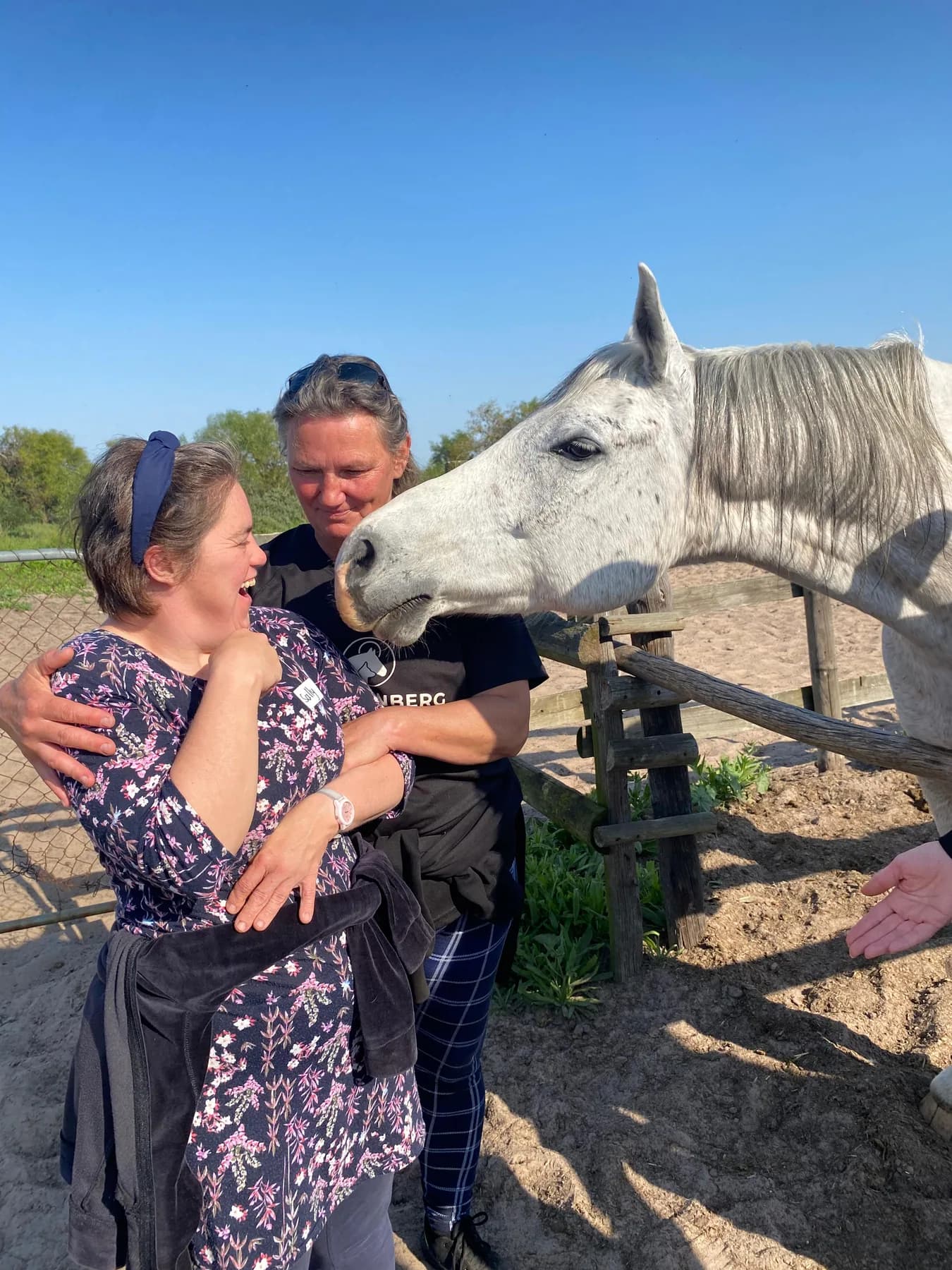 A resident laughing with delight beside a grey horse leaning over the paddock rail, supported by a therapist with an arm around her shoulders.