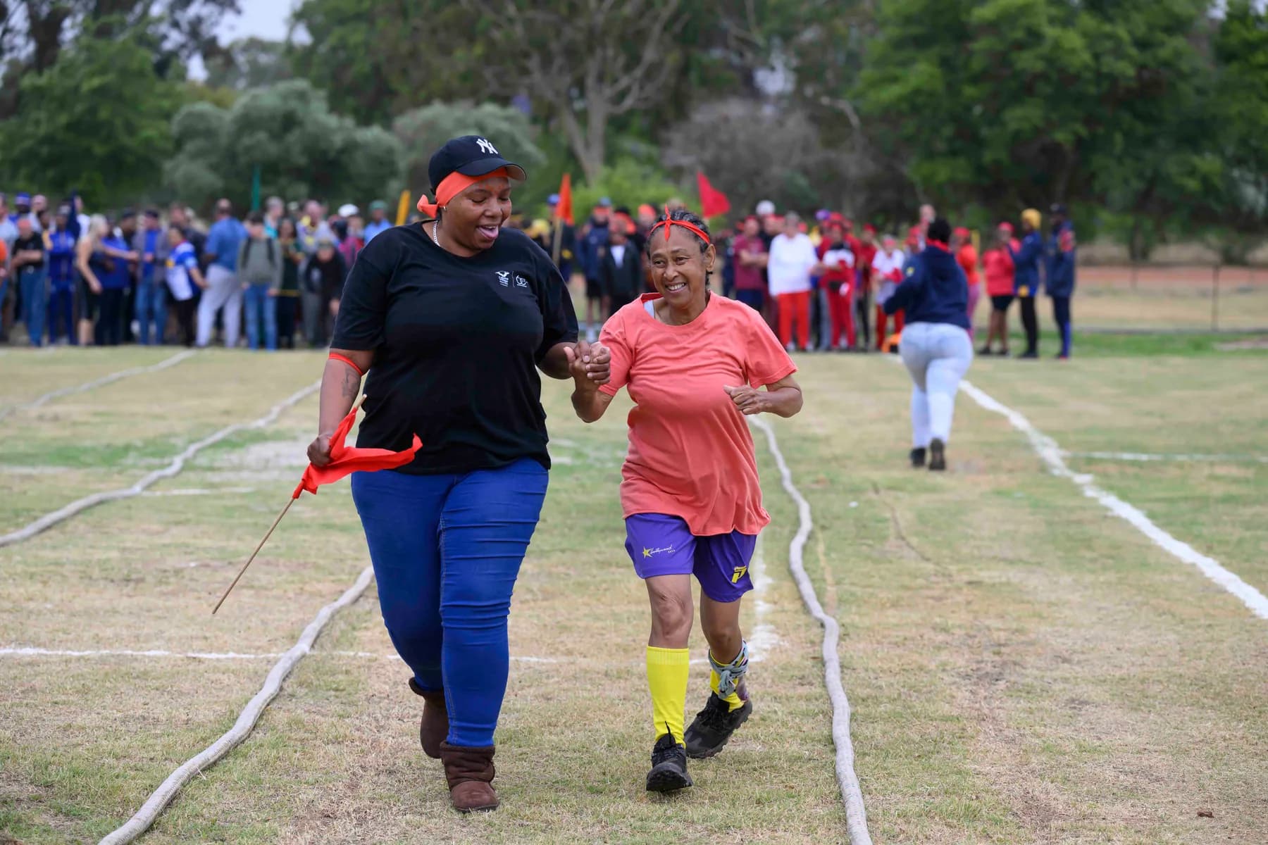 A resident and a staff member running hand in hand down the marked lanes of the sports field while the rest of the village cheers from the sidelines.