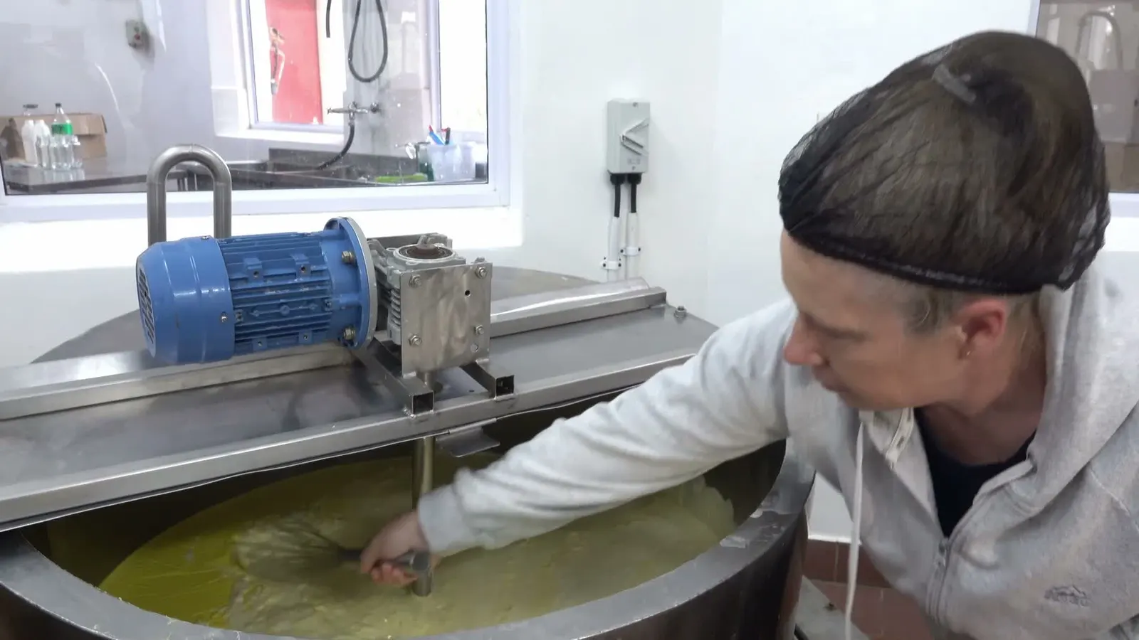 A worker in a hairnet leaning over an open stainless steel vat in the Camphill dairy, reaching in beside the motorised stirrer.