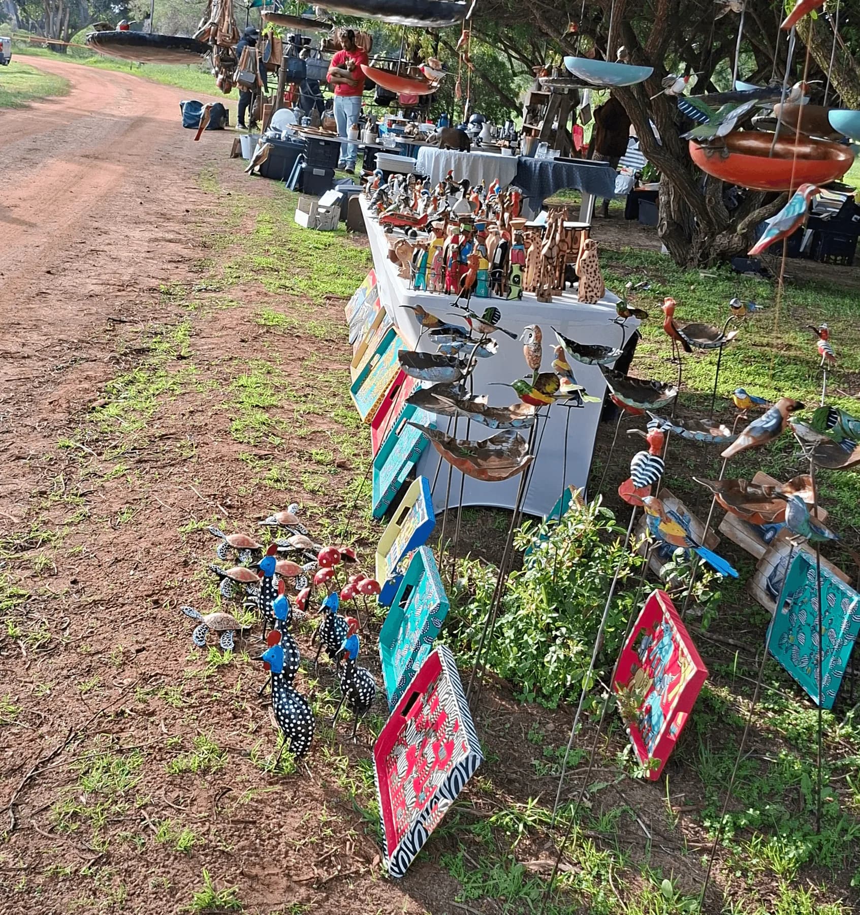 Hand-painted wooden bird sculptures on stakes and brightly painted craft panels lining a sandy farm track beside the market stalls