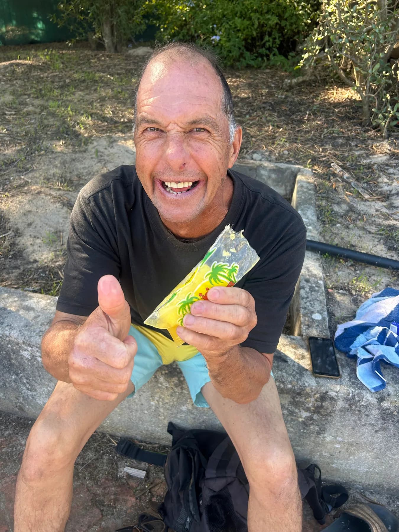 A resident sitting on a rock in the sun, grinning broadly and giving a double thumbs up while holding up a yellow packet.