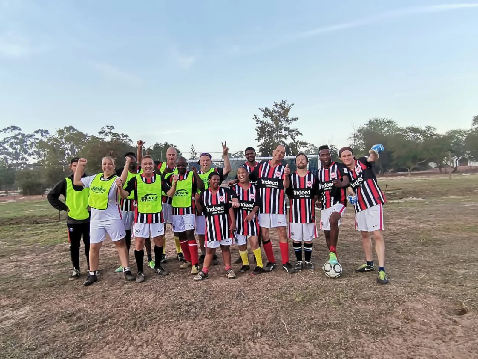 The village soccer team lined up on the field in red and black striped kit and green training bibs, cheering with a ball at their feet.