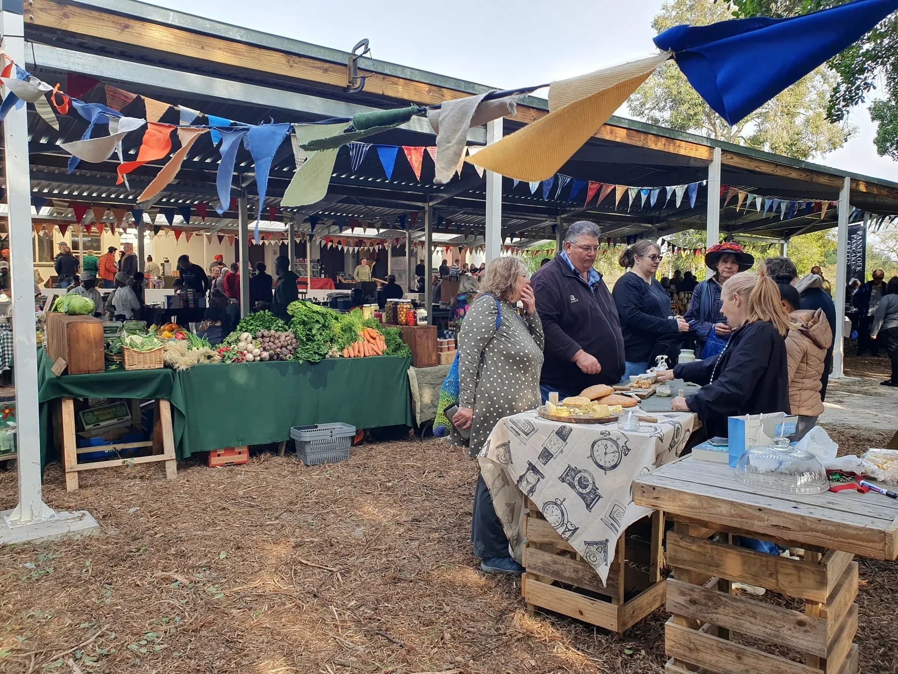 Visitors browsing vegetable and craft stalls under bunting and shady trees at the Camphill monthly market