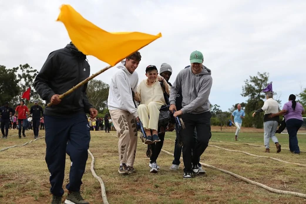 A team carrying a resident on a stretcher during a sports day relay, led by a marshal waving a yellow flag