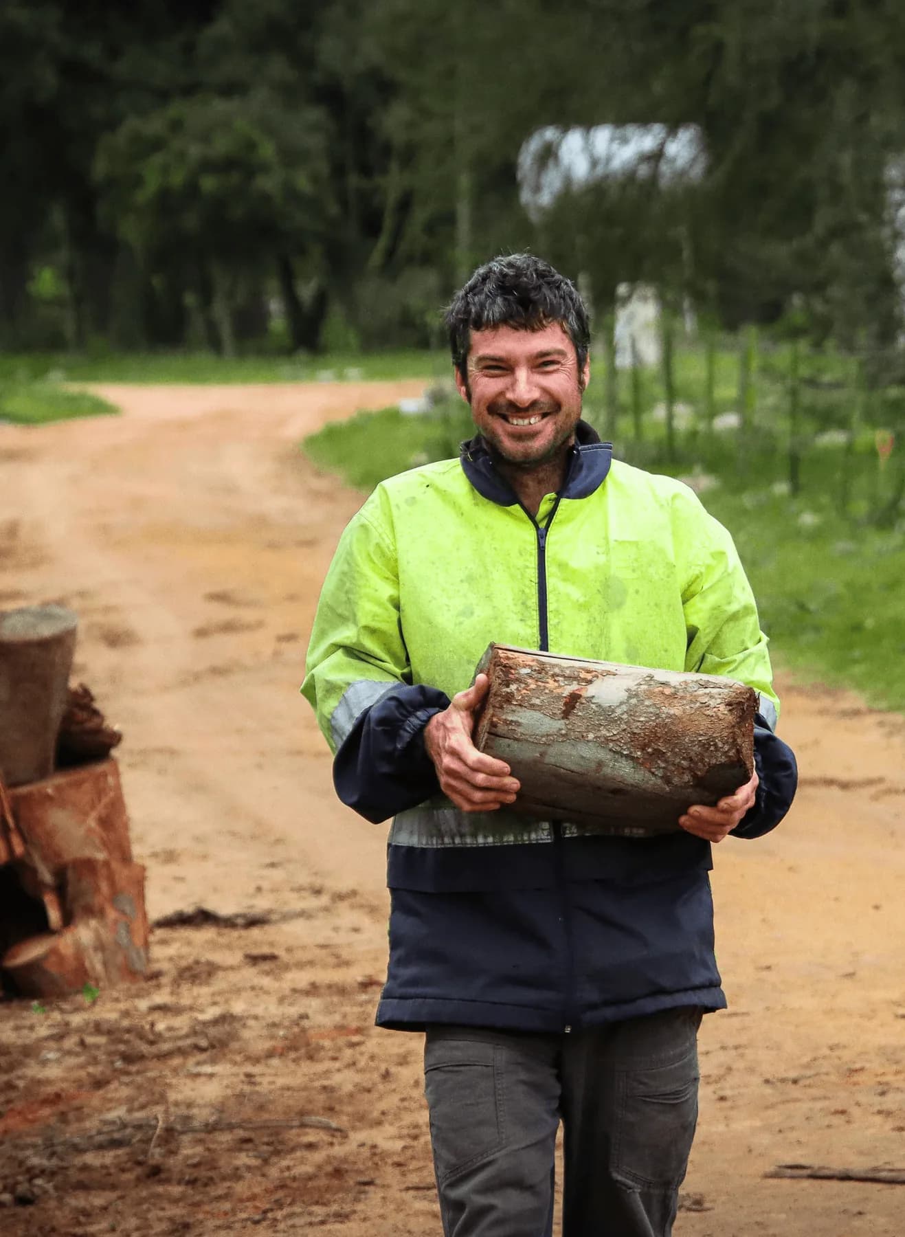 A Camphill worker in a high-visibility jacket carrying a log along a sandy farm road